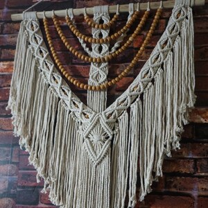 May include: A white macrame wall hanging with wooden beads. The hanging is in the shape of a triangle with a diamond pattern in the center. The hanging has long fringe on the bottom.