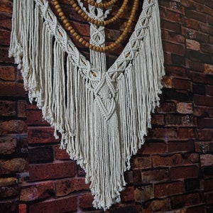 May include: A white macrame wall hanging with a wooden bead garland. The macrame is in a V-shape with fringe on the bottom.