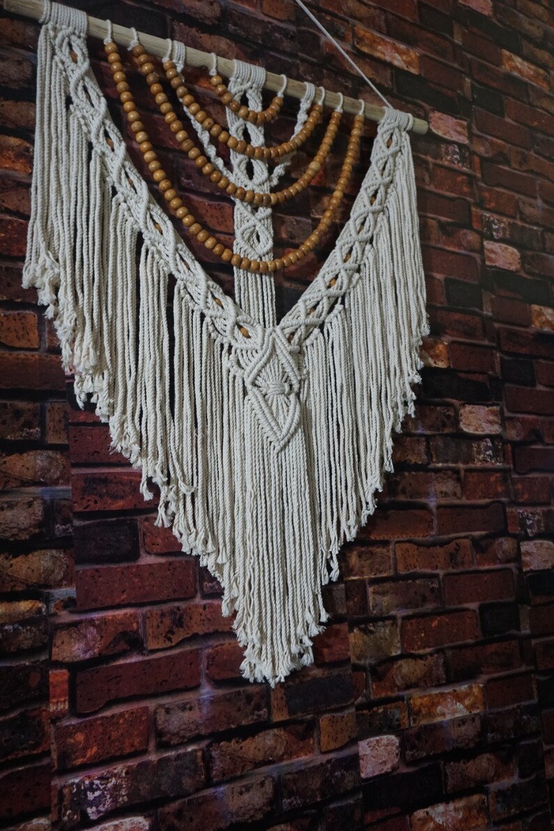 May include: A white macrame wall hanging with a wooden bead garland. The macrame is in a V-shape with fringe on the bottom.