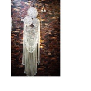 May include: A white macrame dream catcher with five circular woven panels hanging from a brick wall. The dream catcher has long white fringe hanging from the bottom.