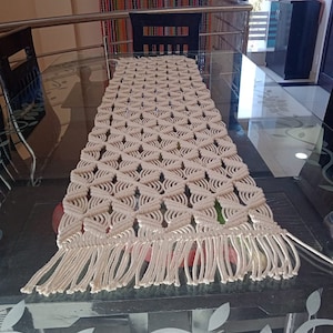 May include: A white crocheted table runner with a geometric pattern and fringe. The runner is on a glass table with a floral design.