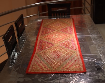 May include: A red table runner with gold and pink embroidery. The runner features a geometric pattern and is made from a soft, textured fabric.