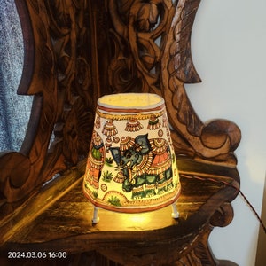 May include: A small, decorative lamp with a painted elephant design. The lamp has a white shade with colorful illustrations and rests on three white legs. The lamp is lit, casting a warm glow. The background features a carved wooden surface.