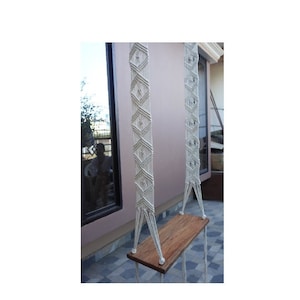 Puede incluir: Una hamaca de macramé blanca con un asiento de madera. La hamaca está colgada del techo de un patio.