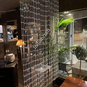 May include: A clear beaded curtain hangs from the ceiling in a restaurant. The beads are strung on a metal rod and are about 1 inch in diameter. The curtain is about 8 feet tall and 4 feet wide. The curtain is in front of a window with a view of a street.
