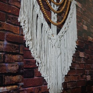 May include: A white macrame wall hanging with wooden beads. The macrame is in a triangular shape with fringe on the bottom. The wooden beads are in a layered pattern.