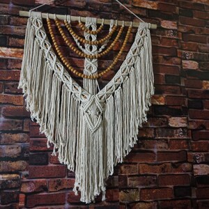 May include: A white macrame wall hanging with wooden beads. The wall hanging is in the shape of a teardrop and has a fringe around the bottom.