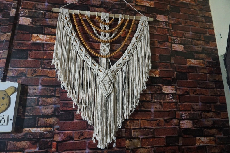 May include: A white macrame wall hanging with wooden beads. The wall hanging is in the shape of a teardrop and has a fringe around the bottom.