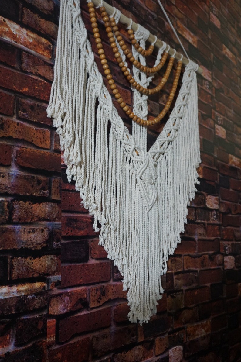May include: A white macrame wall hanging with wooden beads. The hanging is in the shape of a teardrop and has a fringe border.