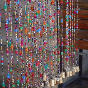 Op de afbeelding: Een kleurrijk kralen gordijn met kleine bellen die aan de onderkant hangen. De kralen zijn in tinten rood, blauw, groen, oranje en geel. De bellen zijn zilver en hebben een glanzende afwerking.