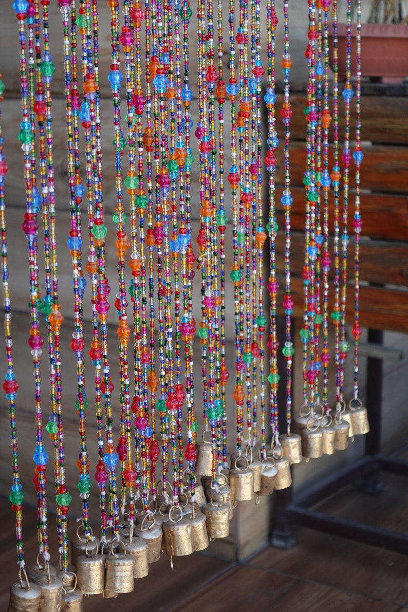 Op de afbeelding: Een kleurrijk kralen gordijn met kleine bellen die aan de onderkant hangen. De kralen zijn in tinten rood, blauw, groen, oranje en geel. De bellen zijn zilver en hebben een glanzende afwerking.
