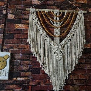 May include: A white macrame wall hanging with wooden beads. The hanging is in the shape of a triangle with a diamond pattern in the center. The hanging is attached to a wooden dowel rod.