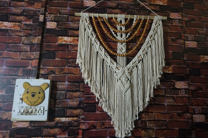 May include: A white macrame wall hanging with wooden beads. The hanging is in the shape of a triangle with a diamond pattern in the center. The hanging is attached to a wooden dowel rod.