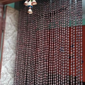 May include: A brown wooden beaded curtain hangs in a doorway. The beads are round and have a dark brown colour. The curtain is made of many strands of beads, and it is hanging from a wooden frame.