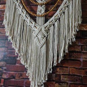 May include: A white macrame wall hanging with wooden beads. The hanging is in the shape of a teardrop and has a fringe border. The wooden beads are arranged in a symmetrical pattern.