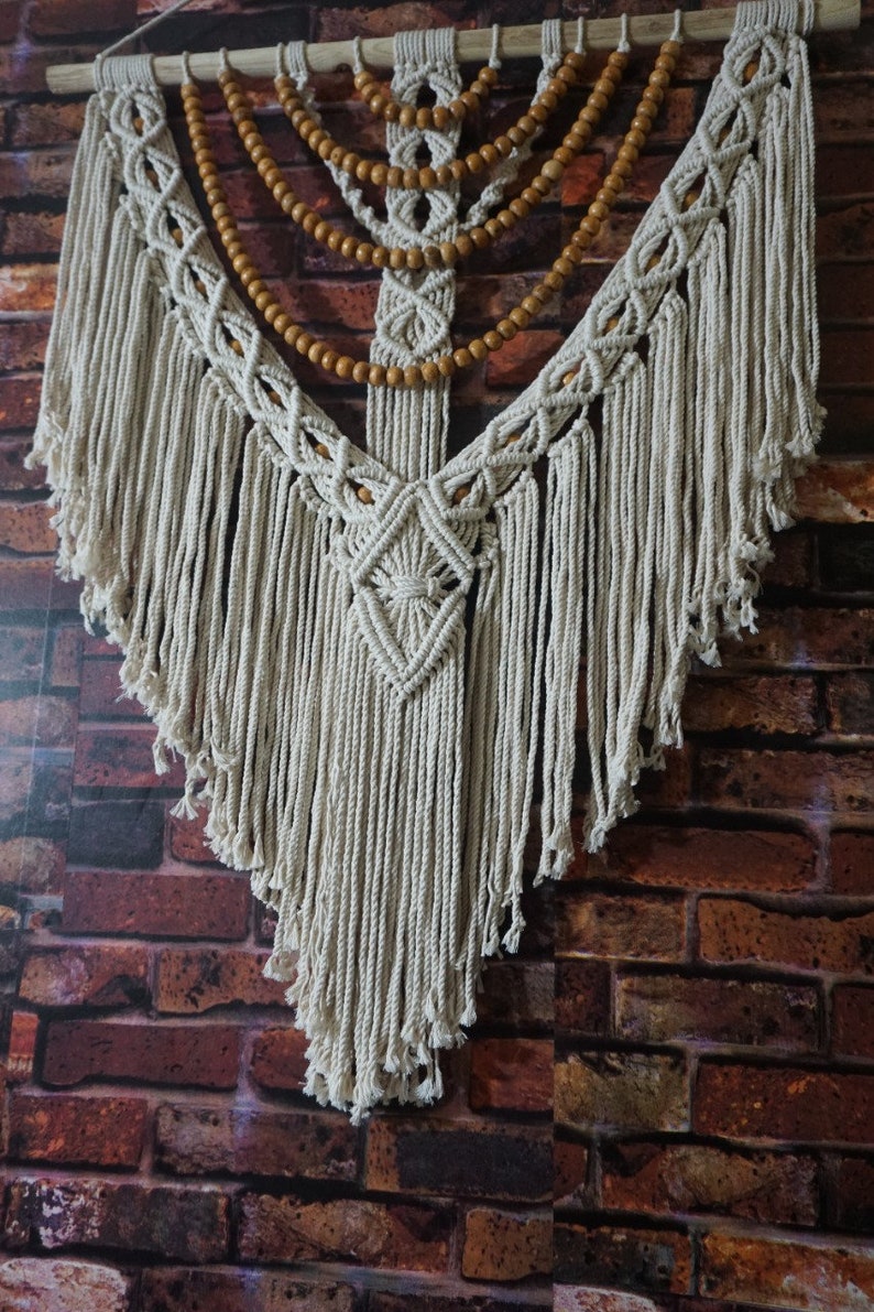 May include: A white macrame wall hanging with wooden beads. The hanging is in the shape of a teardrop and has a fringe border. The wooden beads are arranged in a symmetrical pattern.