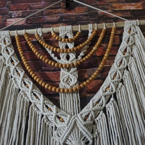 May include: A white macrame wall hanging with wooden beads. The macrame is in a diamond pattern with fringe hanging from the bottom. The wooden beads are strung in a curved pattern across the top of the macrame.