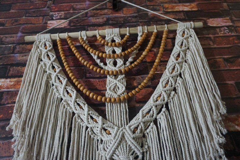 May include: A white macrame wall hanging with wooden beads. The macrame is in a diamond pattern with fringe hanging from the bottom. The wooden beads are strung in a curved pattern across the top of the macrame.