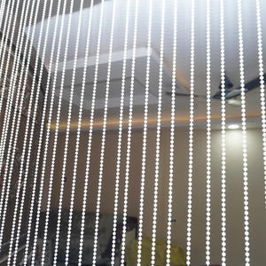 Può includere: Tenda di perline bianche appesa in una porta. La tenda è composta da numerosi fili di piccole perline bianche rotonde. Le perline sono distanziate in modo uniforme, creando una barriera semitrasparente.