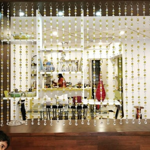 May include: A room divider made of clear and yellow beaded strands hangs in front of a kitchen. The beads are strung on thin wires and are suspended from a metal rod. The kitchen is visible through the beads, and there are shelves and cabinets in the background.