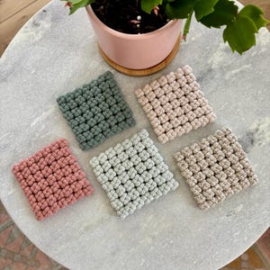 May include: Four square crocheted coasters in different colors: green, pink, light blue, and beige. The coasters are made with a textured, bumpy design.
