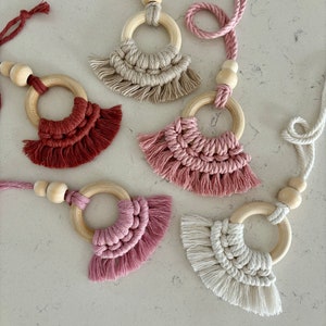 May include: Five macrame keychains with wooden rings and tassels. The keychains are in shades of beige, pink, and red.
