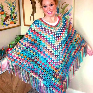 May include: A vibrant, hand-crocheted poncho featuring a square pattern in shades of blue, orange, pink, and green. The poncho has a fringed hem and a round neckline. The garment is worn over a white top.