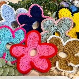 May include: A collection of colorful, handmade crochet flower hair accessories in various shades of blue, pink, yellow, and brown. Each flower has a central opening for hair. The flowers are arranged on a white tray with greenery.
