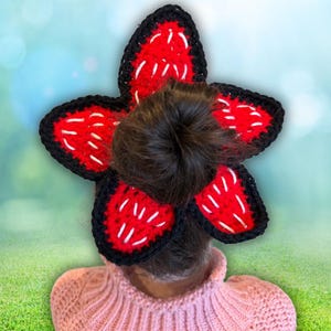 May include: A handmade, crocheted Demogorgon flower hair accessory. The petals are red with white accents and black trim. The accessory is worn on top of a bun, with a pink sweater in the foreground. The background is a blurred green and blue.