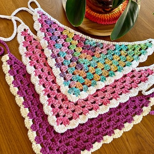 May include: Three triangular crochet bandanas featuring a granny square pattern. One bandana is multicolored with pastel tones, one is solid pink, and one is solid purple. Each piece is finished with a white scalloped border and white ties.