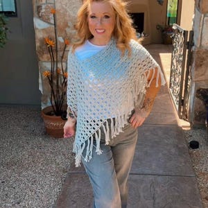 May include: A light gray crochet poncho with fringe detail. The poncho is worn over a white top and paired with gray pants and sandals. The image is taken outdoors.