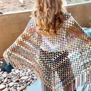 May include: A colorful, open-weave shawl with a fringed hem. The shawl features a mix of colors including red, yellow, and brown. The person is wearing black pants and sandals. The background shows a natural outdoor setting.