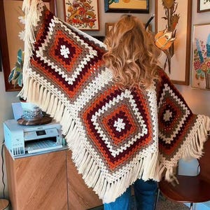 May include: A crochet shawl featuring large square motifs in shades of cream, orange, brown, and dark red. The garment is edged with long cream-colored fringe along the bottom and sides, creating a textured, layered appearance with geometric patterns.