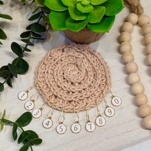 May include: A circular beige crochet piece with eleven gold-toned metal stitch markers attached to the edge. Each marker features a small white circular charm printed with a black number ranging from zero to nine, with an extra one included.
