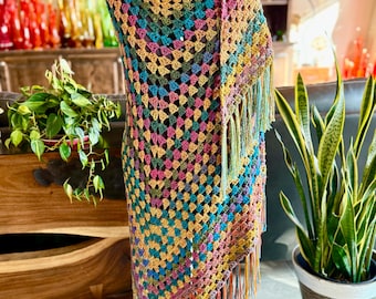 Può includere: Uno scialle triangolare all'uncinetto con un motivo a piastrelle granny multicolore nelle tonalità ottanio, giallo senape, magenta e verde scuro. Il capo è rifinito con lunghe frange sottili e multicolori lungo il bordo inferiore.