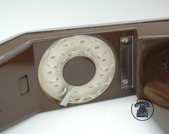 May include: A brown rotary dial phone with a clear plastic dial and a white dial indicator. The phone has a speaker on the side and a cord on the back.