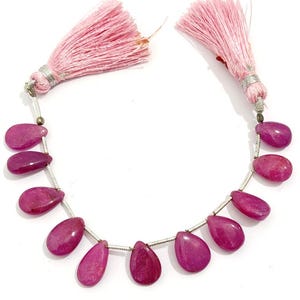 May include: A strand of teardrop-shaped magenta gemstone beads strung on a silver-toned wire, featuring a light pink tassel at each end. The beads are smooth and polished, arranged in a curved line with small silver spacers between each stone.