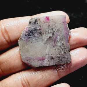 May include: A rough, white and grey gemstone with pink and red streaks. The gemstone is a raw ruby.