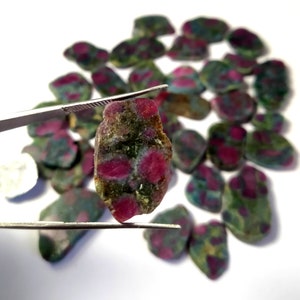 May include: A selection of rough ruby gemstones, displaying a mix of red and green colours. One stone is held by tweezers, highlighting the vivid red spots within the green matrix. The gemstones are scattered on a white surface.
