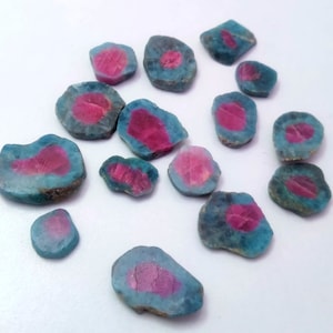 Könnte beinhalten: Eine Sammlung roher, ungeschliffener Edelsteine. Jeder Stein hat einen leuchtend rosa Kern, der von einer teal-blauen Außenschicht umgeben ist. Die Steine variieren in Größe und Form und haben eine raue, natürliche Textur.