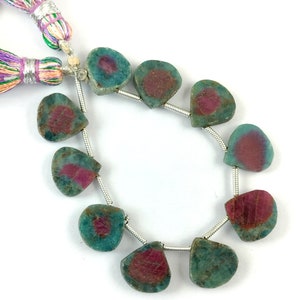 Pode incluir: Fio de contas de pedras preciosas. Cada conta tem uma forma única, com uma camada externa verde-azulado e um centro vermelho escuro. As contas são enfiadas em um fio de prata, com uma borla colorida na extremidade. As pedras preciosas têm uma aparência natural e orgânica.