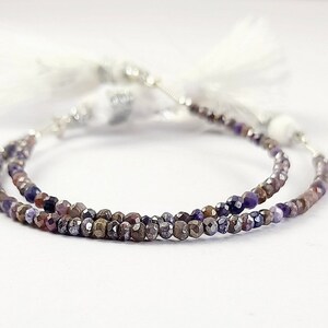 May include: A necklace with small, faceted beads in shades of purple, brown, and silver. The beads are strung together, creating a delicate strand. White tassels are attached to the ends of the necklace.