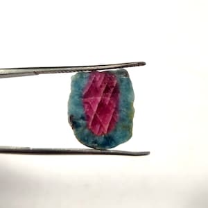 May include: A pink and blue gemstone with a faceted design. The gemstone is held in tweezers.