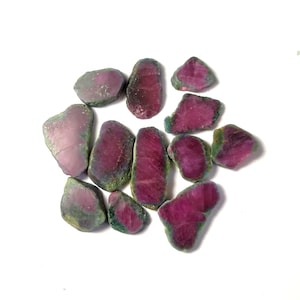 May include: A collection of 11 rough, uncut ruby gemstones. The stones are a deep red color with green inclusions. They are irregularly shaped and have a natural, unpolished finish.