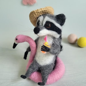 May include: A needle-felted raccoon figurine wearing a tan crocheted hat, sitting inside a pink flamingo-shaped pool float. The raccoon has grey and white fur, black eye patches, and holds a small orange drink with a pink straw in its paw.