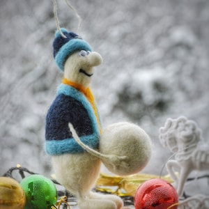May include: A white felt snowman wearing a blue and orange hat and jacket, holding a white felt snowball. The snowman is standing on a wooden base with red and green Christmas ornaments around it.