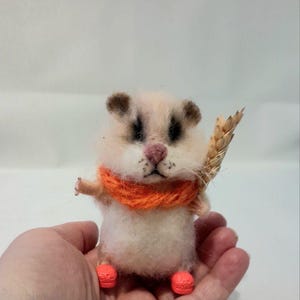 May include: A small, white, needle-felted hamster wearing an orange scarf and orange shoes, holding a stalk of wheat.