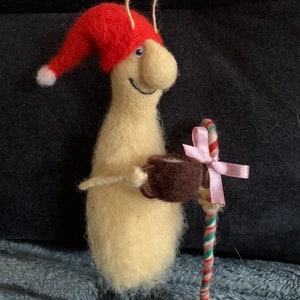 May include: A whimsical felt gnome wearing a red Santa hat, holding a brown mug and a candy cane with a pink bow. The gnome is standing on a wooden base.