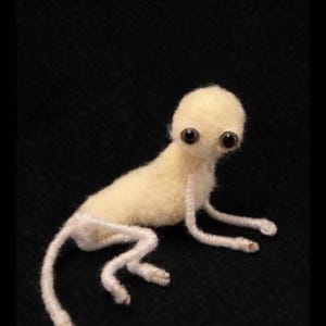 May include: A handmade, light yellow creature with large, dark eyes and a small body. The creature is lying down on a black surface, with its arms and legs extended. The creature is made of a soft, fuzzy material.