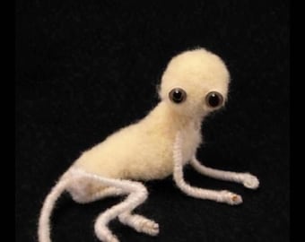 May include: A handcrafted, pale yellow creature with large, dark eyes and a small body. The creature is lying on a black surface, with its arms and legs extended. The creature is made of a soft, fuzzy material.